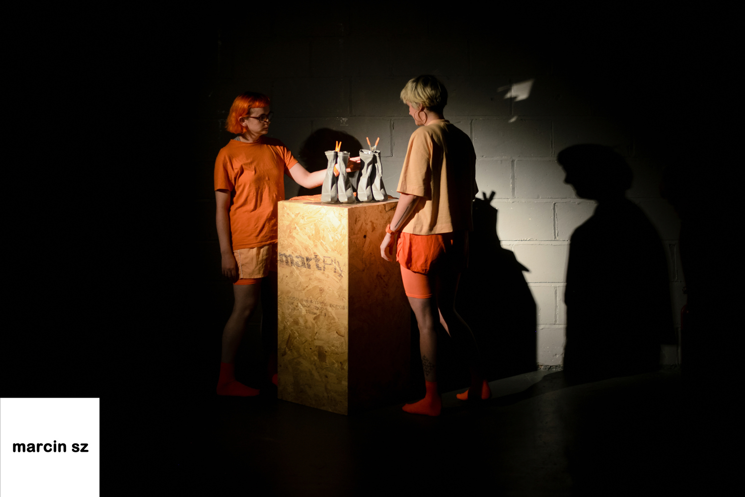 Emily and Emily stand in a spotlight next to a wooden plinth. On it are two pairs of reflective heeled boots.