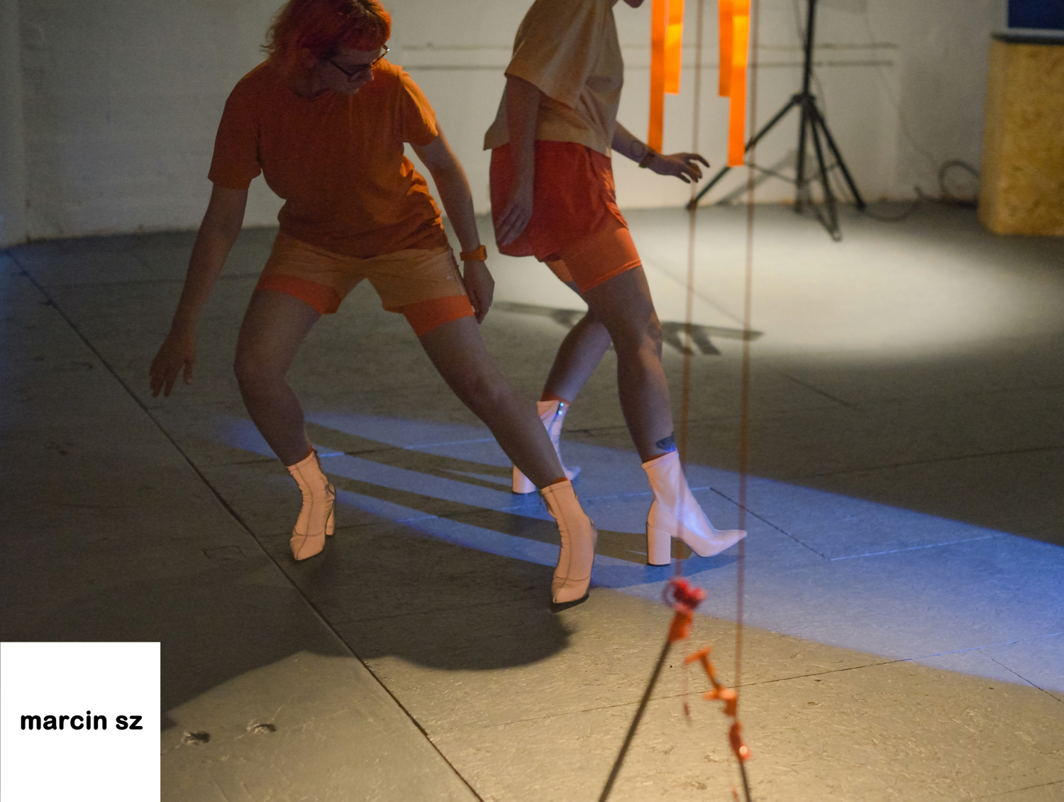 Emily and Emily stand back to back, wearing all orange in an open gallery space. They both have one leg stretched out to the side, wearing reflective heeled boots.