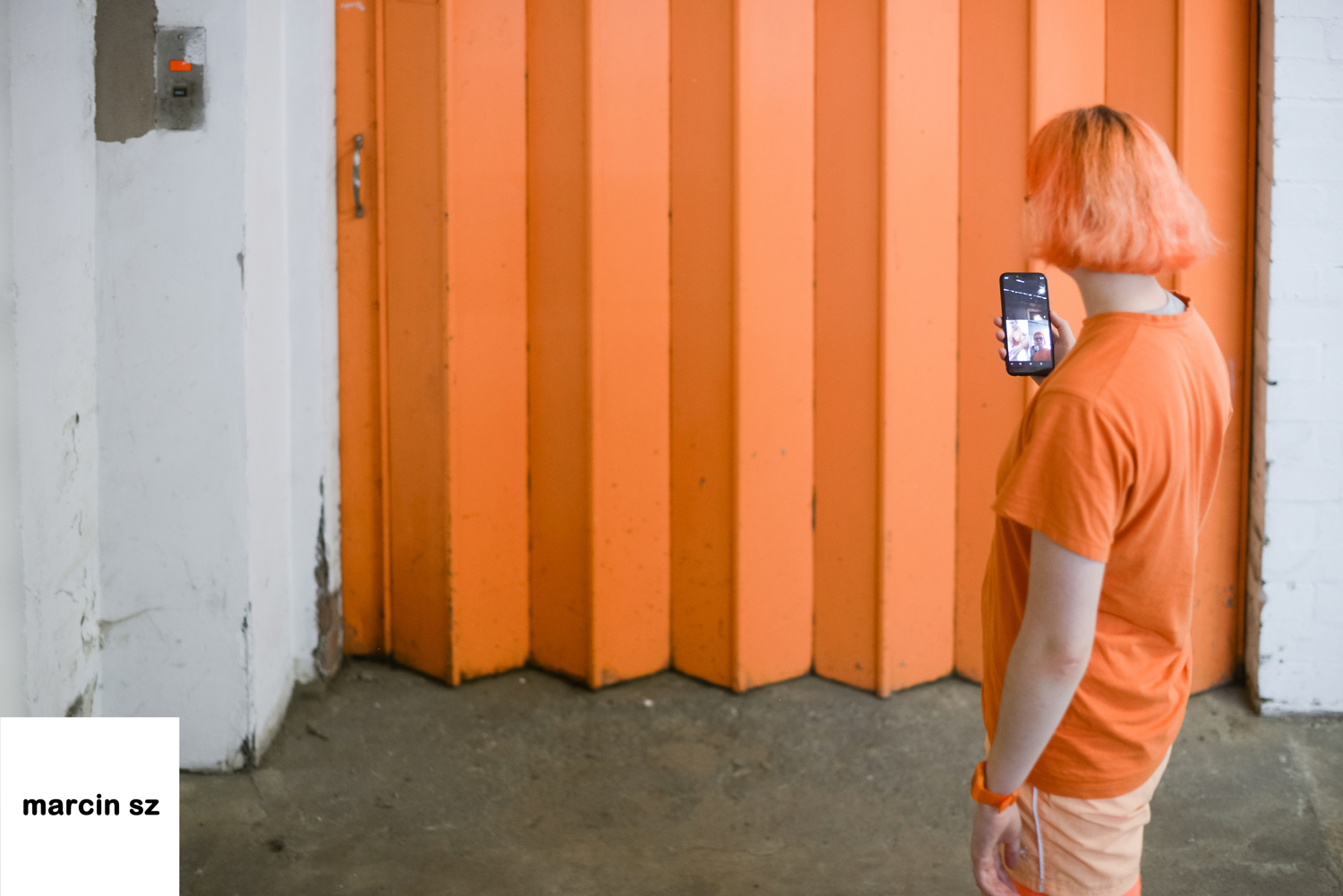 Emily R faces away from the camera, dressed in orange in front of a large orange shutter door. She looks at a phone screen on a call.